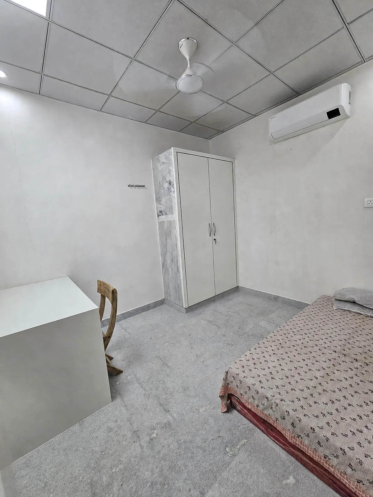 Wider view of the representative furnished room showing the bed, wardrobe, desk, chair, AC and ceiling fan